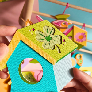 Cabane en papier découpé à la main avec un trèfle à quatre feuilles sur le toit, assortie au Messager du cœur n°3. Un abri porte-bonheur, fabriqué à partir de 12 couches de papiers découpés, dans les teintes orange, vert et bleu.