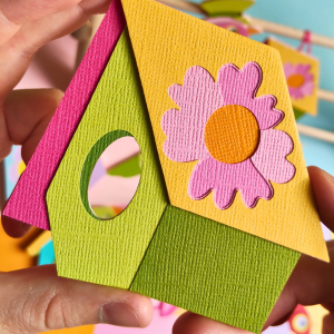 Cabane en papier découpé à la main avec une pâquerette sur le toit, assortie au Messager du cœur n°2. Un abri fleuri, fait de 11 couches de papiers texturés, dans les teintes violet, bleu et vert.