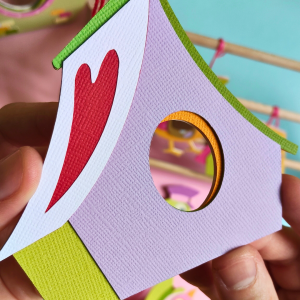 Cabane en papier découpé à la main avec un cœur symbolique sur le toit, assortie à l'oiseau Messager du cœur n°1. Un ornement mural unique, fait de 8 couches de papier, dans les teintes rouge, violet et vert.